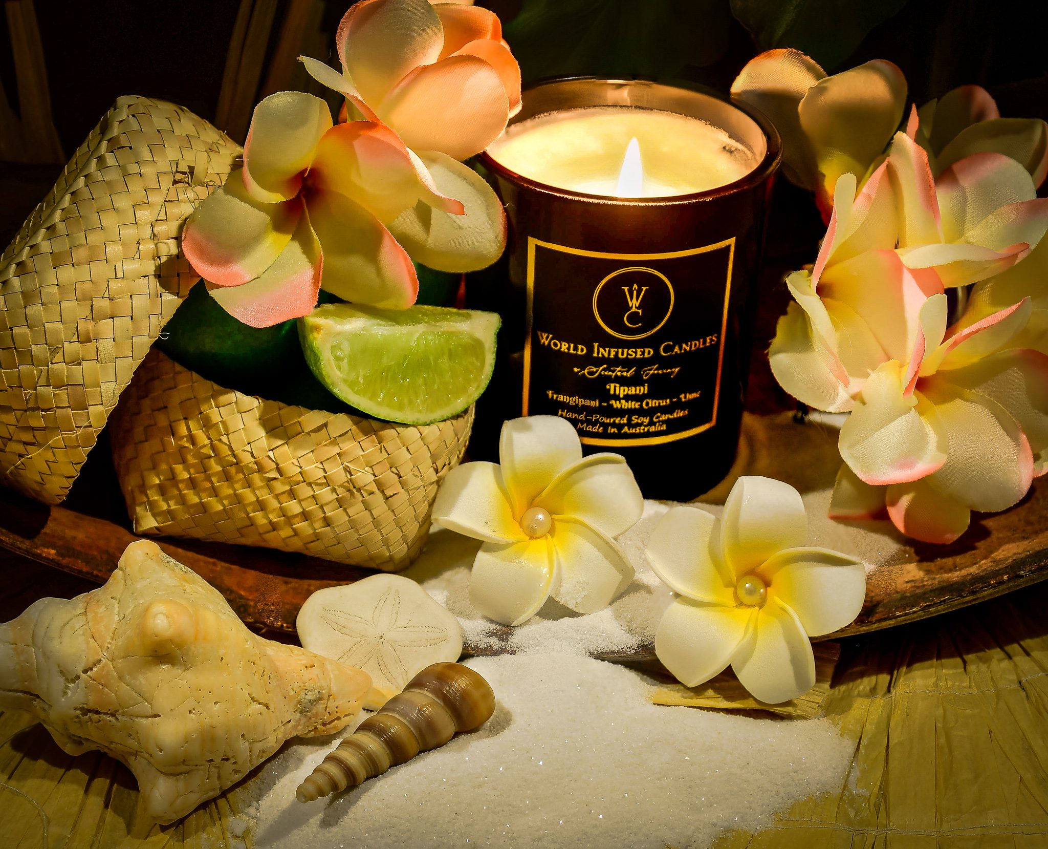 Tipani soy candle example of being lit with sand, shells and frangipani flowers surrounding the candle